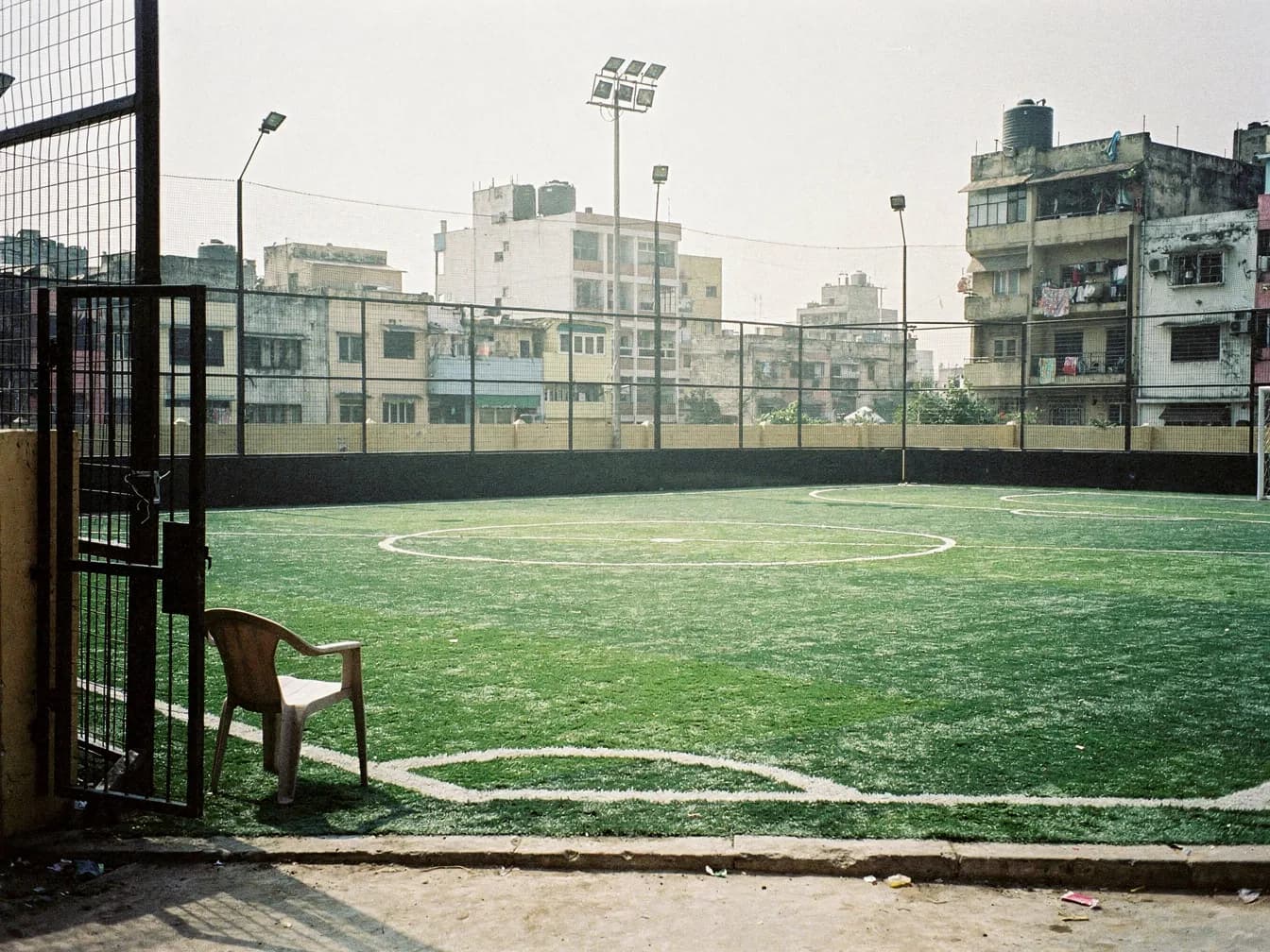An empty five-a-side turf in the afternoon sun, floodlights off and the gate open, a plastic chair by the entrance