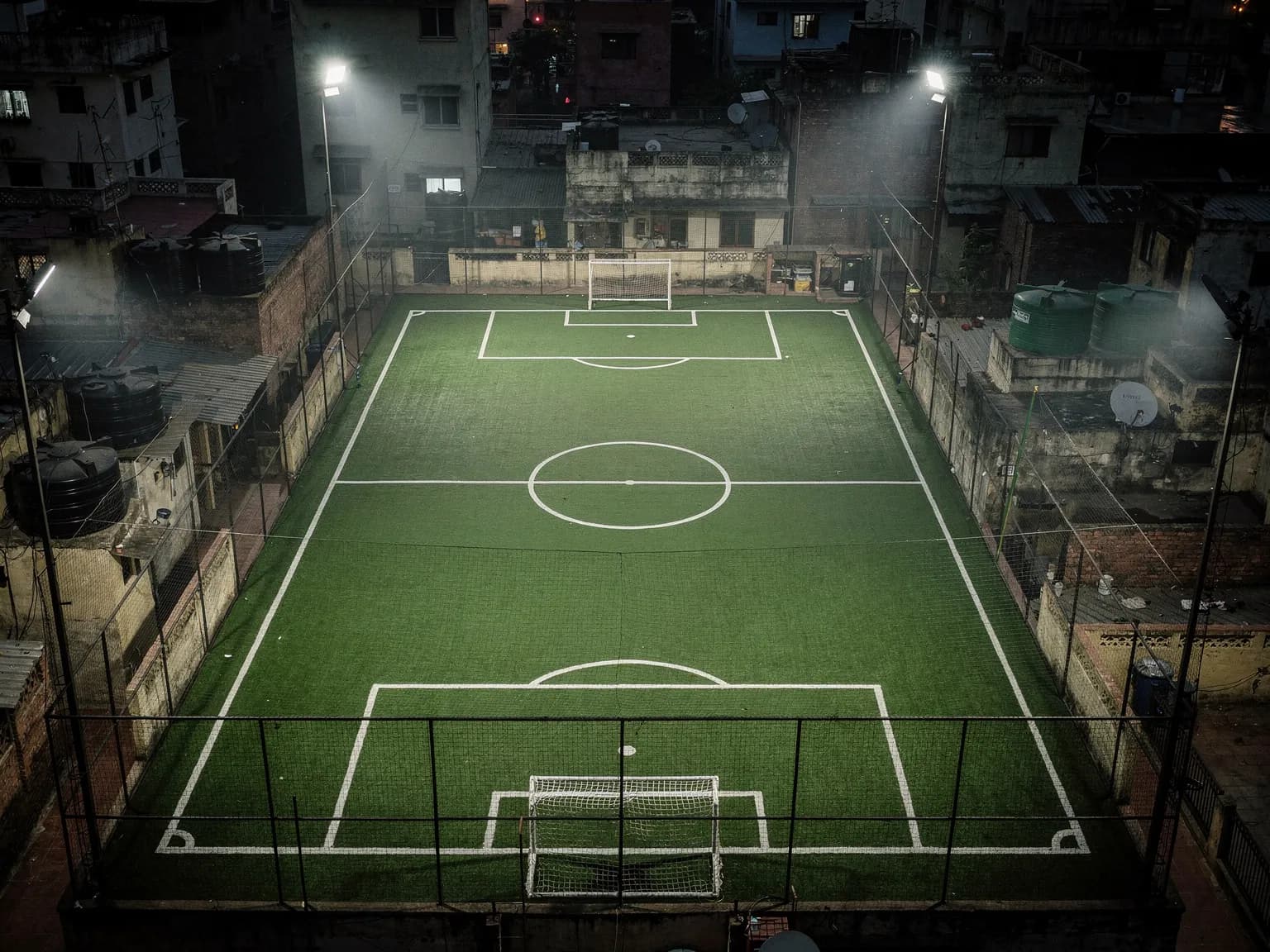 A floodlit rooftop five-a-side turf at night, seen from above among water tanks and rooftops